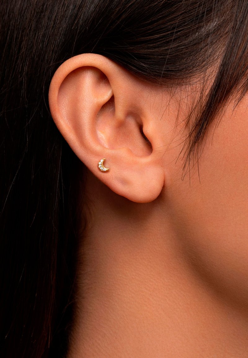 Boucle d'oreille en forme de croissant de lune doré avec de petites pierres transparentes, fixée à une oreille percée. L'oreille a une forme ovale avec une texture de peau lisse.