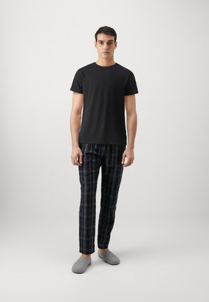 Νεαρός άνδρας που στέκεται φορώντας απλό μαύρο μπλουζάκι, σκουρόχρωμο μπλε καρό παντελόνι και γκρι παντόφλες, μπροστά σε απλό φωτεινό φόντο.