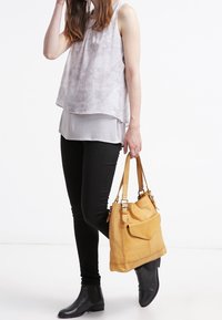 Borsa a tracolla in pelle gialla con tasca frontale. Il modello indossa una maglietta grigia chiara con motivo, jeans skinny neri e stivaletti neri.