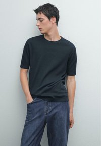 Černé tričko s krátkým rukávem a kulatým výstřihem, vyrobené z hladkého materiálu, v kombinaci s volnými tmavými džínami, které vykazují klasický design a jednoduchou siluetu.