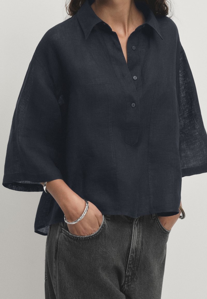 Femme portant une chemise ample en lin bleu marine foncé à manches mi-longues, rentrée dans un jean noir délavé taille haute, mains dans les poches, bracelets argentés aux poignets.