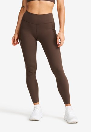 Bruna högmidjade leggings gjorda av elastiskt material, med en mjuk textur och subtila sömnadsdetaljer, kombinerade med vita sneakers.