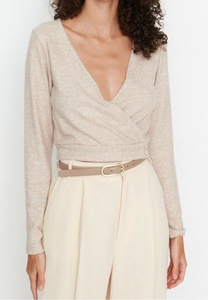 Vrouw die een beige wikkelstijl top met lange mouwen draagt, met een diepe V-hals, gecombineerd met hoog getailleerde crèmekleurige broeken en een beige riem.