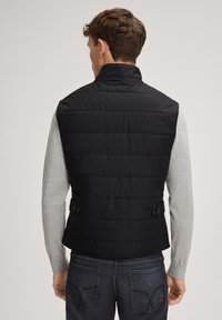 Czarna pikowana kamizelka z wysokim kołnierzem, z przeszywanym wzorem i ozdobami w postaci guzików po bokach, noszona na jasnoszarej koszuli z długim rękawem.