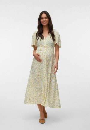 Femme enceinte souriante portant une robe jaune à fleurs avec des manches papillon, tenant son ventre, debout devant un fond blanc uni.