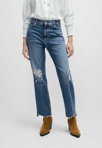 Blå denimjeans med hög midja och rak passform. Har en sliten yta på vänstra knäet och fransad fåll. Bärs med bruna mocka stövlar.
