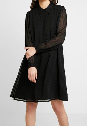 Woman wearing a black long-sleeve dress with textured dots, buttoned front, collar, and sheer sleeves, standing against a plain background.