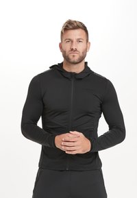 Sort atletisk zip-up jakke med hætte, tætsiddende design, lange ærmer, glat tekstur og lynlåslommer på siderne.