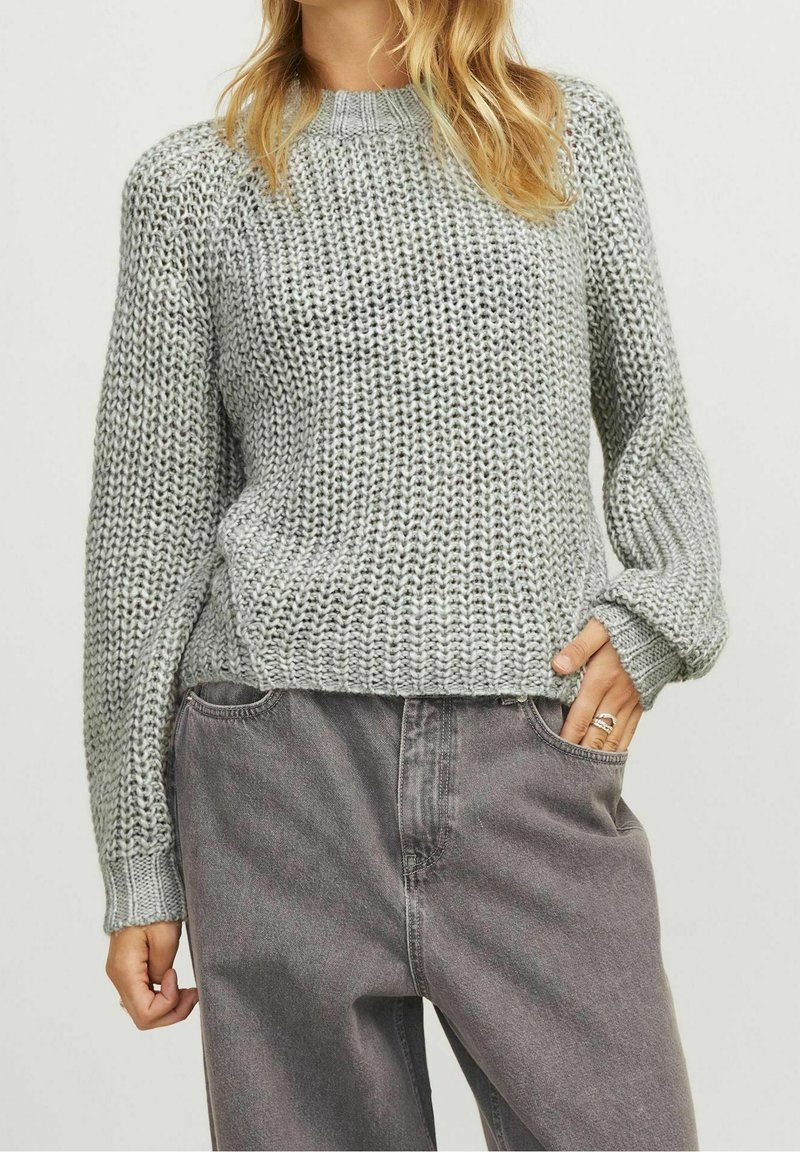 Femme portant un pull gris ample en maille et un jean noir délavé taille haute, une main dans la poche et des bagues visibles aux doigts.