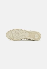 Suela de zapatilla en goma color blanco roto con un patrón de agarre texturizado; presenta elementos de diseño circulares y lineales, con el logo de la marca visible.