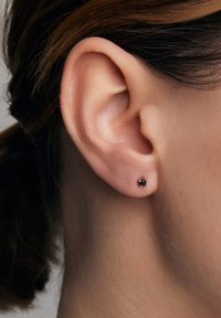 Złoty kolczyk wkręcany z małym czarnym kamieniem w centrum, umieszczony na tle ucha i włosów osoby.