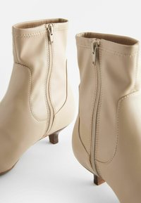 Beige ankle boots with a smooth synthetic material, side zippers, short block heels, and subtle stitching details.