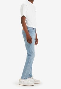 Personne portant un jean en denim bleu clair, un t-shirt blanc à manches courtes, des baskets blanches, debout de profil sur un fond blanc.