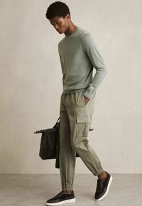 Suéter de manga larga verde claro, pantalones cargo oliva con puños elásticos y zapatillas oscuras. El modelo lleva una bolsa negra. Look suave y casual.