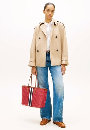 Vrouw draagt een beige dubbelreeks jasje, wit overhemd, blauwe spijkerbroek, bruine schoenen, houdt een rode tas met gestreept ontwerp vast.