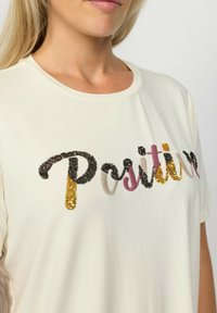 Cremefarvet bomulds T-shirt med korte ærmer og rund halsudskæring, der har multikolor "Positive" broderi i palietter og trådstrukturer.