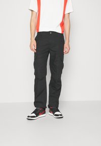 Svarta cargobyxor med flera fickor, i kombination med en vit t-shirt med röda vertikala ränder och svarta sneakers med röda accenter.