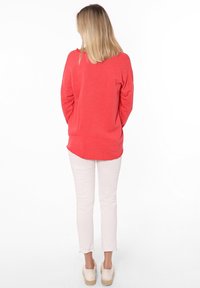 Maglietta a maniche lunghe color corallo con orlo arrotondato, abbinata a pantaloni aderenti di colore chiaro e scarpe espadrillas beige. Tessuto morbido e vestibilità rilassata.