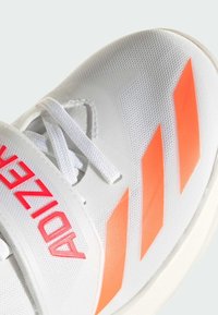 Baskets blanches avec un dessus texturé, présentant trois rayures oranges et une sangle avec un logo rose. Les lacets sont visibles sur le dessus.