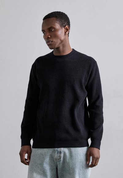 Pull en maille côtelée noir avec un col rond et des manches longues, associé à un jean en denim bleu clair. Design simple et texturé.