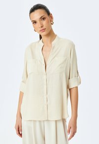 Koszulka w kolorze kremowym z kołnierzykiem mandaryńskim, dwoma przednimi kieszeniami i podwiniętymi rękawami. Wykonana z lekkiego, teksturowanego materiału.
