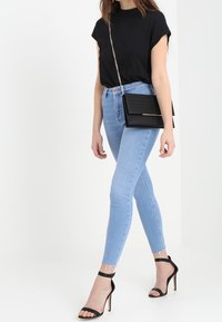 Kvinna som bär en svart kortärmad topp, ljusblå skinny jeans, svarta ankelremssandaler och bär en svart kedjeryggsäck.