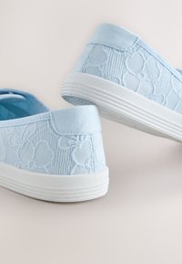 Une paire de chaussures bleu clair à enfiler avec des semelles blanches et une broderie florale texturée sur la tige en tissu, présentées vues de dos et de côté.