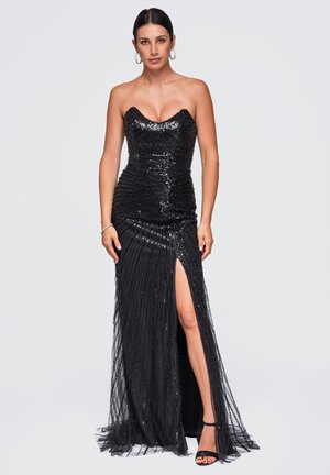 Femme portant une robe noire sans bretelles pailletée avec une fente haute sur la cuisse, des talons noirs, des boucles d'oreilles anneaux argentés et un bracelet.