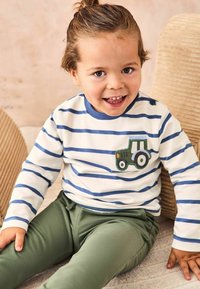 Gestreiftes langärmliges Hemd in Weiß und Marineblau, mit einer grünen Traktor-Applikation auf der Brust; kombiniert mit olivgrünen Hosen.