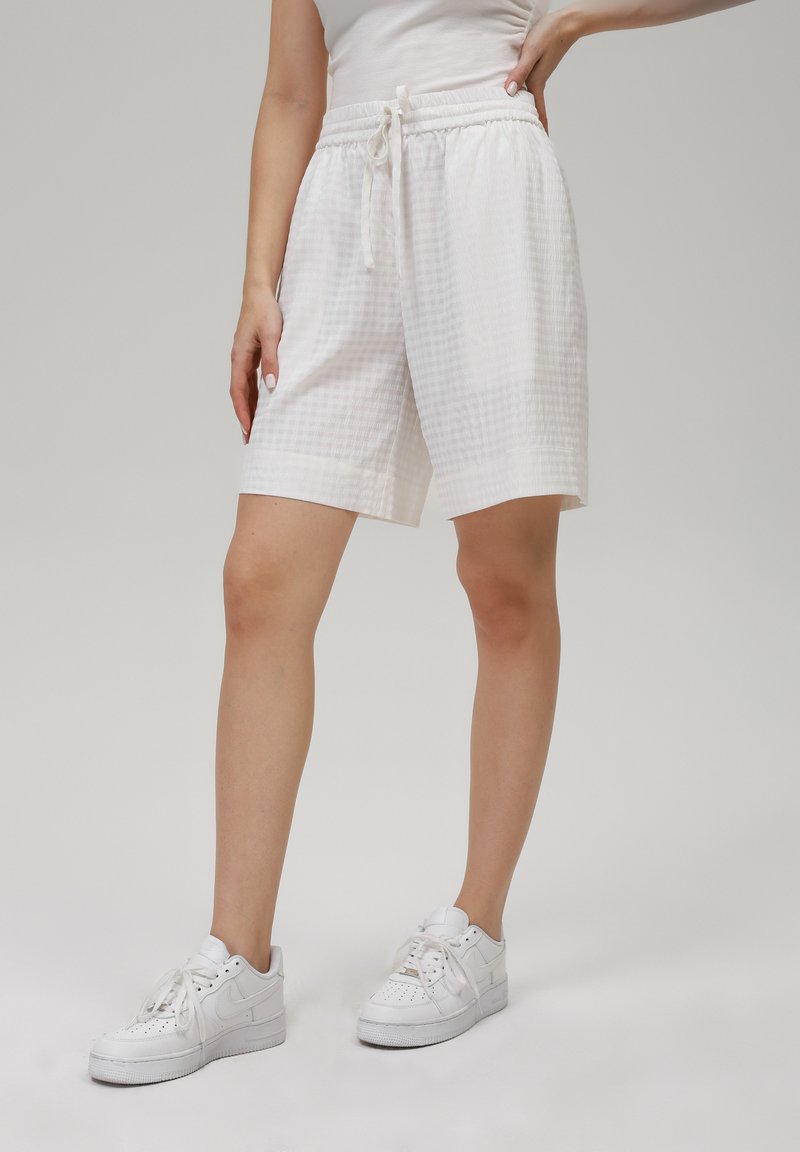 Witte, gestructureerde shorts met een elastische tailleband en trekkoorddetail, met een lengte tot halverwege de dij en een relaxte pasvorm. Gedragen met witte sneakers.