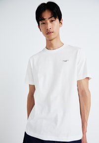 Witte katoenen T-shirt met korte mouwen, ronde hals en een kleine merkprint aan de linkerkant, met een gladde textuur.