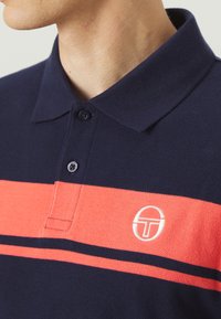 Námornícka polo košeľa s korálovými horizontálnymi prúžkami a bielym logom. Má golier a trojici gombíkov, textúrovaná tkanina.