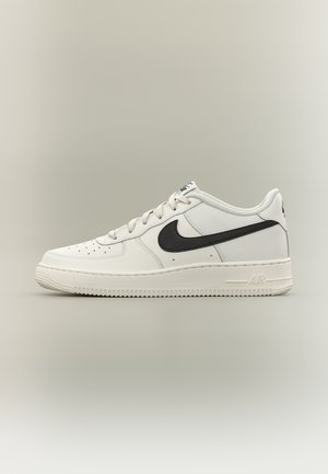 Baskets Nike Air Force 1 basses blanches avec logo Swoosh noir, laçage à l'avant et boîte à orteils perforée sur fond neutre.