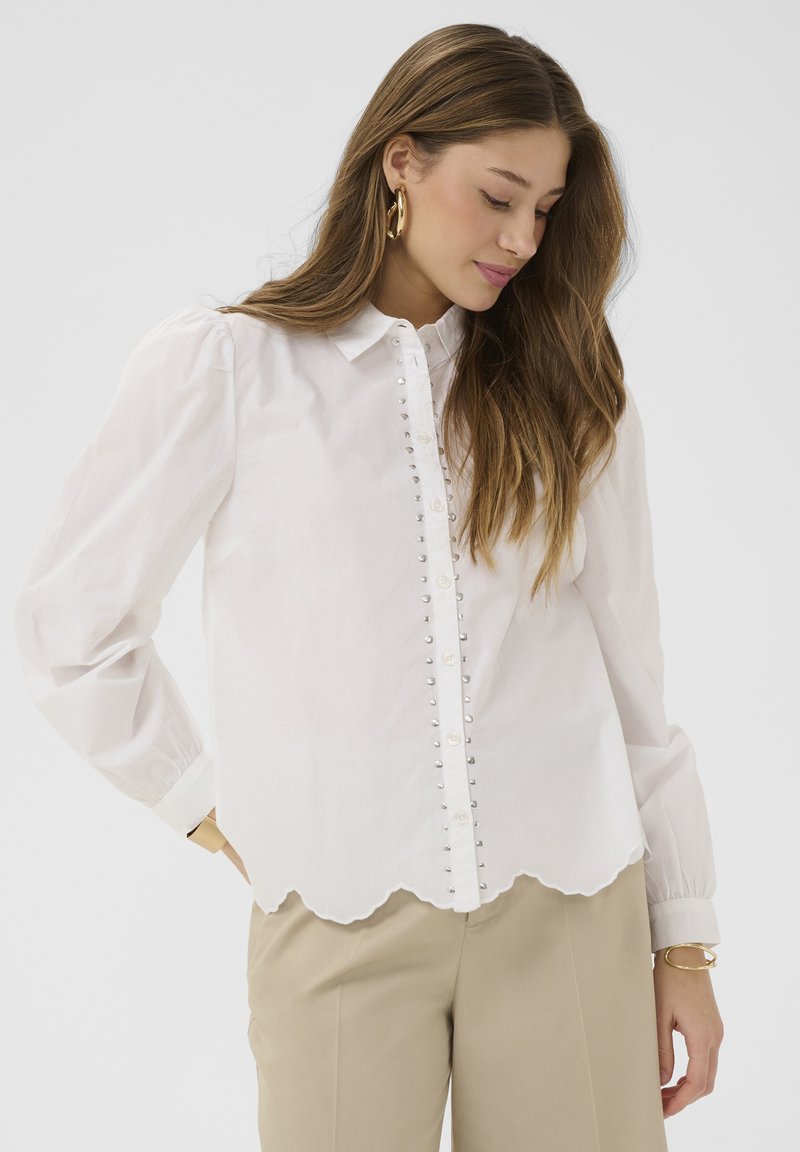 Femme aux longs cheveux bruns portant des boucles d'oreilles créoles dorées, un chemisier blanc à boutons avec un ourlet festonné et un pantalon beige, regardant vers le bas avec réflexion.