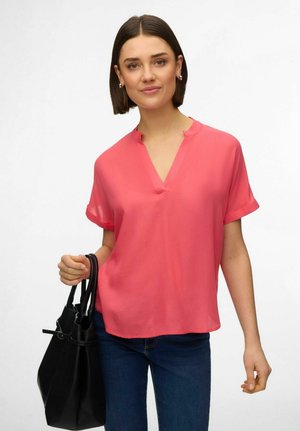 Femme aux cheveux courts bruns portant un chemisier rose corail et un jean sombre, tenant un sac à main noir sur un fond blanc.