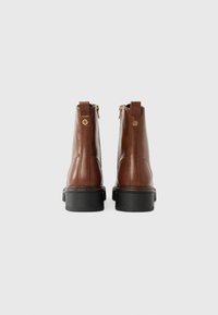 Bottines en cuir marron avec deux zips dorés, une semelle noire texturée et une finition lisse. La vue arrière montre un léger talon et des accents de logo.