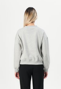 Kvinna med långt blont hår som vänder sig bort, iklädd en ljusgrå sweatshirt och svarta byxor mot en enkel vit bakgrund.