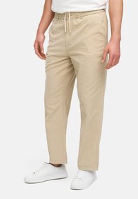 Pantalones de mezcla de algodón beige con cintura elástica y cordón, que cuentan con bolsillos laterales y un diseño de pierna recta. Llevados con zapatillas blancas.