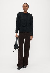 Γυναίκα που φοράει μαύρη υφασμάτινη ζακέτα, σκούρο παντελόνι με φαρδιά μπατζάκια, μαύρα παπούτσια πλατφόρμα και κρατάει μια μικρή μαύρη τσάντα.