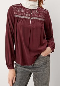 Frau in einer burgunderfarbenen Langarmbluse mit Spitzenverzierungen und silbernen Knöpfen über einem weißen Rollkragenpullover, kombiniert mit grauen Jeans.