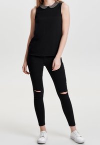 Top nero senza maniche con un colletto decorativo, abbinato a jeans skinny neri strappati e sneakers bianche. La texture è liscia e aderente.
