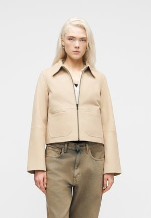 Blondína so svetlou pokožkou v béžovej bunde na zips a vybledlých olivovo zelených džínsach stojaca pred jednoduchým svetlým pozadím.