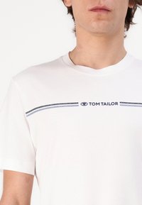 Hvit bomull t-skjorte med rund hals, korte ermer og en horisontal marineblå stripe med "TOM TAILOR" trykket i midten.