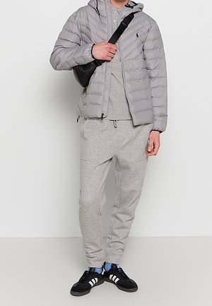 Mężczyzna ubrany w szarą kurtkę puchową, szare polo, szare dresy, czarną torbę na ramię oraz czarne buty sportowe Adidas z białymi paskami.