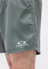 Πράσινα σορτς Oakley στο μηρό ενός ατόμου με ορατή ραφή πλαϊνής τσέπης και λογότυπο της μάρκας κοντά στο τελείωμα.