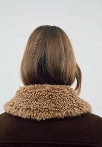 Brun uldfrakke med en tekstureret, beige faux pelskrave. Kraven er fyldig og rammer bagsiden af nakken og håret.