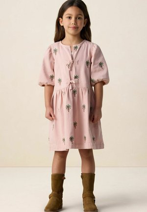 Jeune fille portant une robe rose à imprimé de palmiers et des bottes marron mi-mollet, debout devant un fond beige uni.