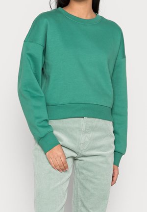Sweatshirt vert court avec un col rond, des poignets et un ourlet côtelés. Le matériau semble doux avec une texture lisse. Associé à un pantalon en velours côtelé vert clair.