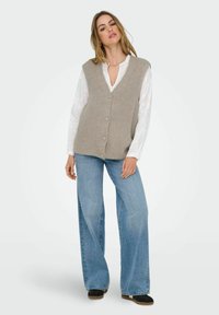 Gilet lavorato a maglia beige con scollatura a V e chiusura con bottoni, abbinato a jeans blu chiaro larghi. Il tessuto è a coste con una finitura testurizzata.