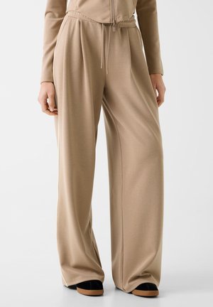 Beżowe szerokie spodnie z elastycznym pasem i sznurkiem, noszone z pasującą beżową bluzką i czarnymi butami z brązowymi podeszwami.